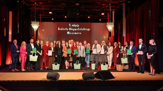 Grupa osób z dyplomami i zielonymi teczkami na scenie przed ekranem z napisem o siódmej edycji Budżetu Obywatelskiego Mazowsza.