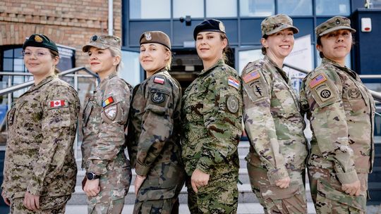 Sześć kobiet w mundurach z naszywkami Kanady, Rumunii, Polski, Chorwacji i USA stoi przed budynkiem.