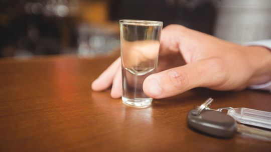Dłoń trzymająca kieliszek z przezroczystym płynem, obok na drewnianym blacie leżą kluczyki samochodowe.