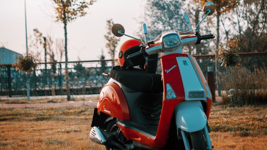 Czerwono-szary skuter Vespa zaparkowany na trawie, z czerwonym kaskiem na siedzeniu.