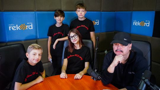 Czworo młodych muzyków w czarnych koszulkach z nazwą Chwilowo Bez Nazwy oraz ich opiekun przy mikrofonach w studiu radiowym.