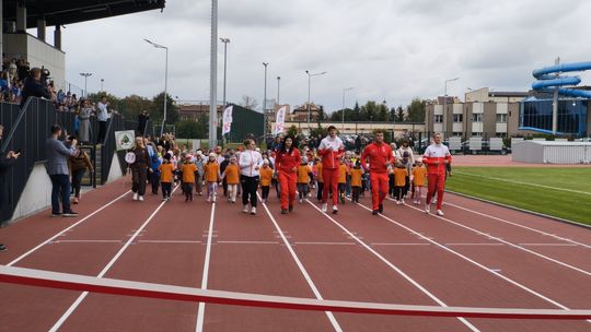 Grupa dzieci w pomarańczowych koszulkach oraz dorośli sportowcy biegną po czerwonej bieżni nowo otwartego stadionu w Płońsku, zbliżając się do czerwonej wstęgi na mecie. Z lewej strony na trybunach widać zgromadzonych kibiców, a w tle zabudowę oraz błękitną zjeżdżalnię.