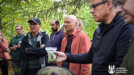 Grupa osób dorosłych w lesie słucha instruktora. Mężczyzna w okularach i czarnej kurtce w pierwszym planie po prawej trzyma mały przedmiot w przezroczystej torbie.