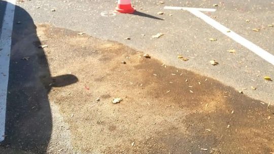 Plama rozlanej cieczy i sorbentu na asfalcie parkingu obok białych linii i pachołka ostrzegawczego.