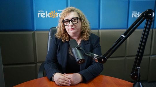 Kobieta z kręconymi blond włosami i w czarnych okularach siedzi przy mikrofonie w studiu Radia Rekord, trzymając dłonie na stole.