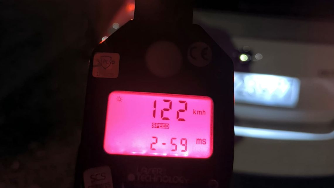 Wyświetlacz miernika prędkości z wynikiem 122 km/h, w tle nieostry tył samochodu.
