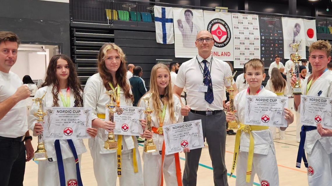 Pięcioro młodych karateków w białych strojach trzyma puchary i dyplomy Nordic Cup. Stoją z dwoma mężczyznami w hali sportowej.