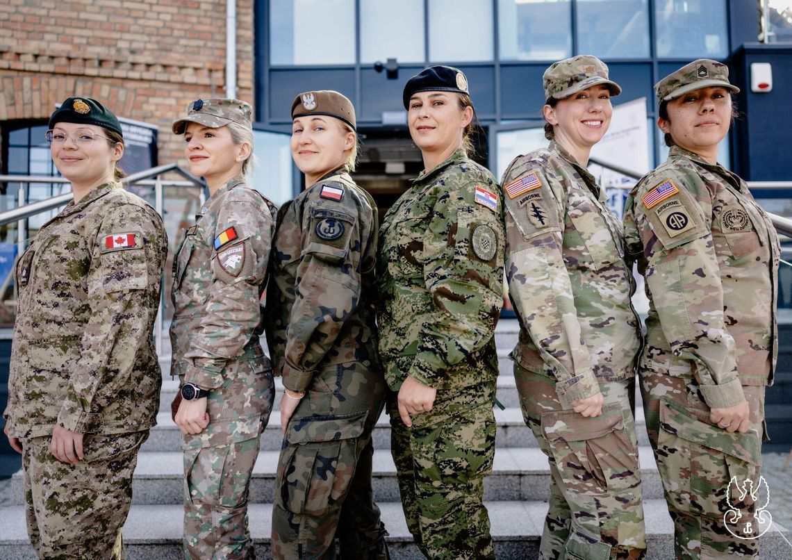 Sześć kobiet w mundurach z naszywkami Kanady, Rumunii, Polski, Chorwacji i USA stoi przed budynkiem.