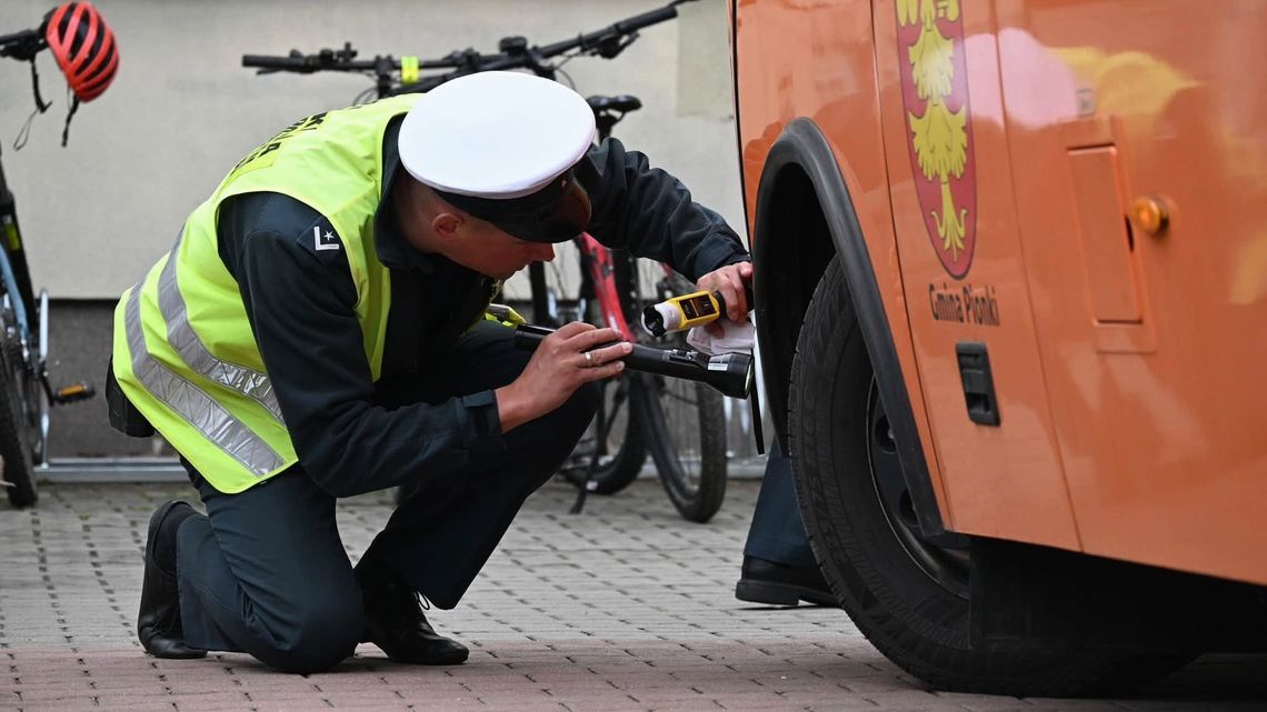 Inspektor Inspekcji Transportu Drogowego w odblaskowej kamizelce i czapce sprawdza latarką stan techniczny koła pomarańczowego autobusu.