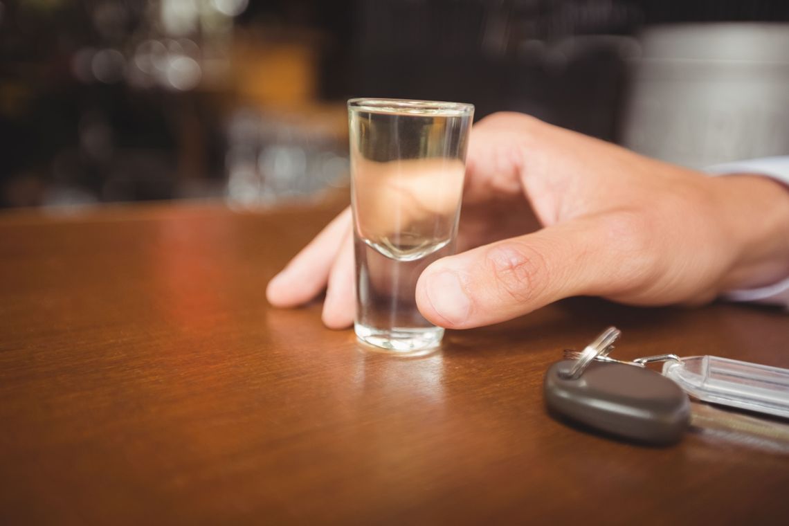 Miał 2,5 promila i zakaz prowadzenia. W gminie Sońsk przesiadł się na tylny fotel Dłoń trzymająca kieliszek z przezroczystym płynem, obok na drewnianym blacie leżą kluczyki samochodowe.