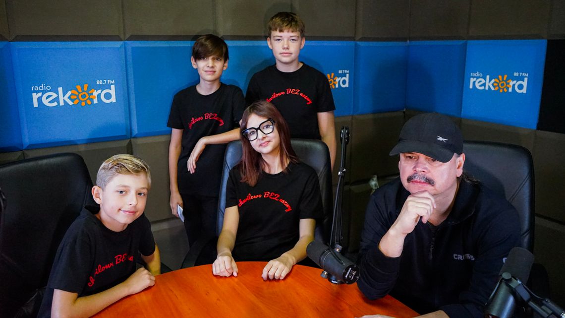 Czworo młodych muzyków w czarnych koszulkach z nazwą Chwilowo Bez Nazwy oraz ich opiekun przy mikrofonach w studiu radiowym.