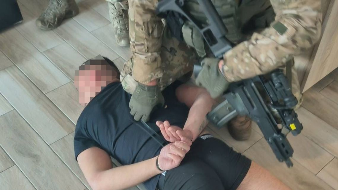 Uzbrojony funkcjonariusz w wyposażeniu taktycznym przy mężczyźnie leżącym na podłodze z rękami skutymi kajdankami.