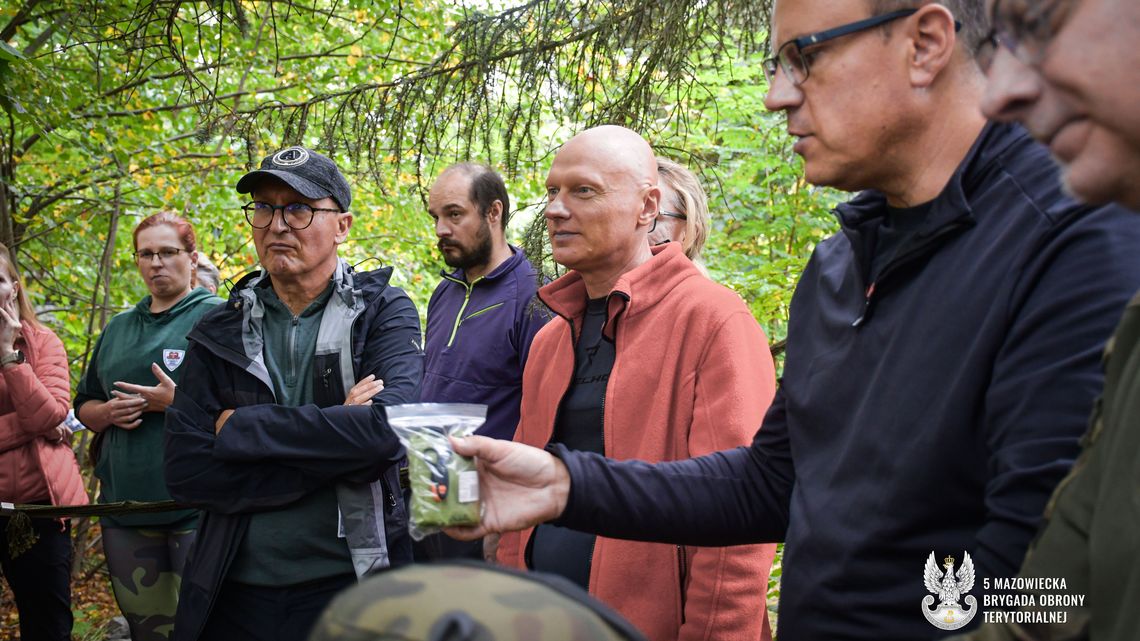 Grupa osób dorosłych w lesie słucha instruktora. Mężczyzna w okularach i czarnej kurtce w pierwszym planie po prawej trzyma mały przedmiot w przezroczystej torbie.