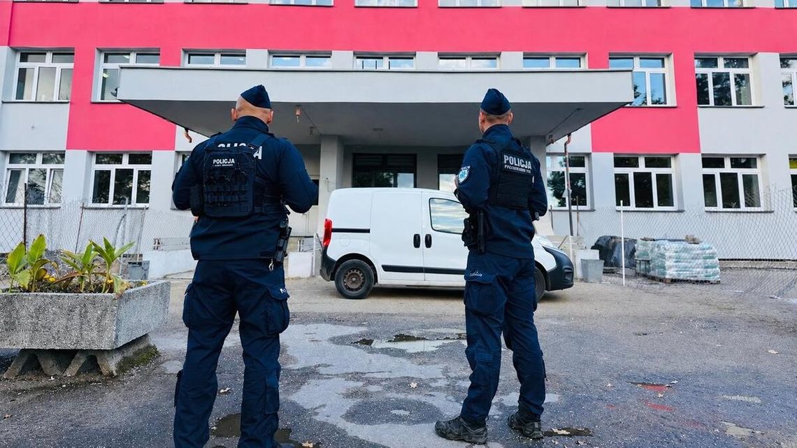 Dwóch umundurowanych policjantów stojących przed wejściem do budynku o biało-czerwonej elewacji.