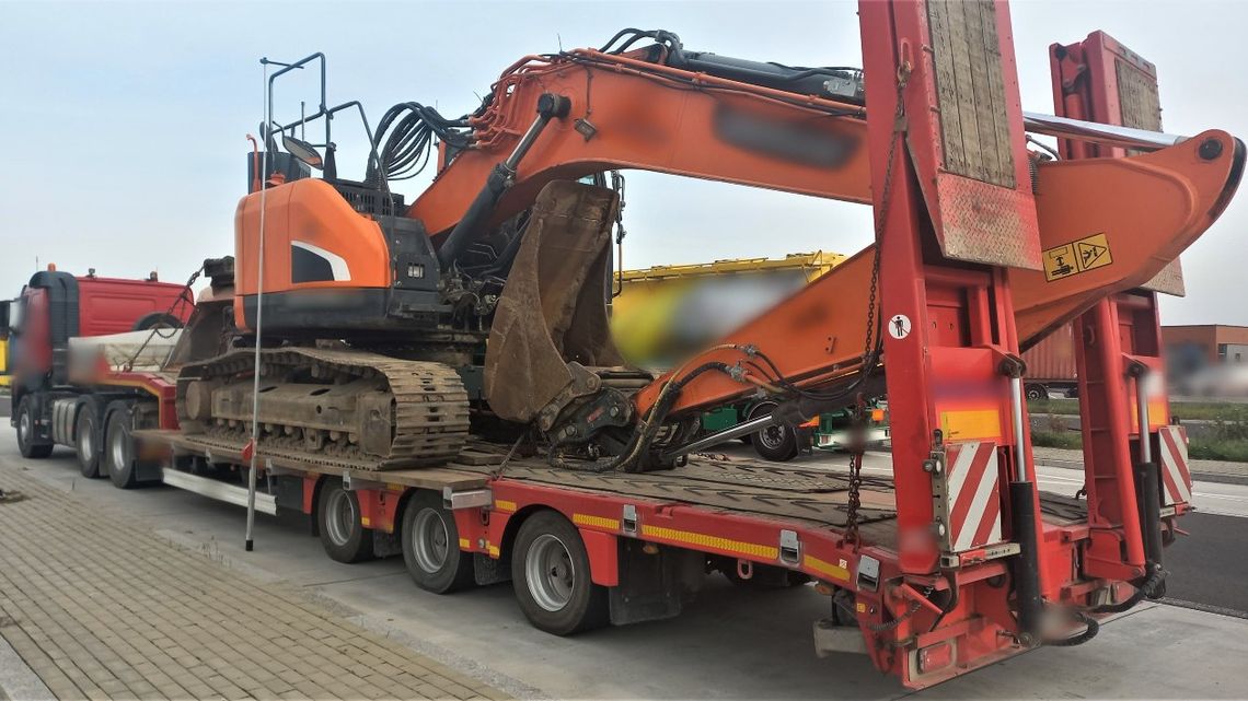Pomarańczowa koparka gąsienicowa z osprzętem na czerwonej naczepie niskopodwoziowej.