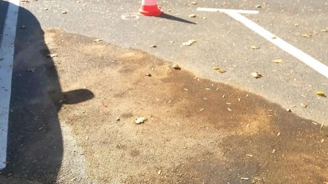 Plama rozlanej cieczy i sorbentu na asfalcie parkingu obok białych linii i pachołka ostrzegawczego.