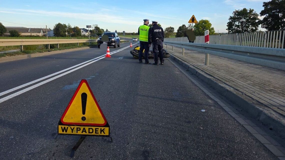Znak ostrzegawczy z napisem wypadek ustawiony na asfalcie na wiadukcie, w tle dwaj policjanci stojący przy przewróconym motocyklu obok barier ochronnych oraz radiowóz