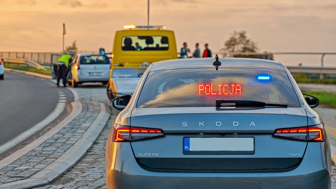 Tylna szyba policyjnej Skody Superb z czerwonym napisem "POLICJA" i włączonymi sygnałami świetlnymi.