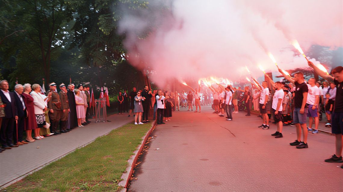 Mieszkańcy, kibice z odpalonymi racami i żołnierze zgromadzeni przed pomnikiem Powstańców Warszawskich w Ciechanowie.